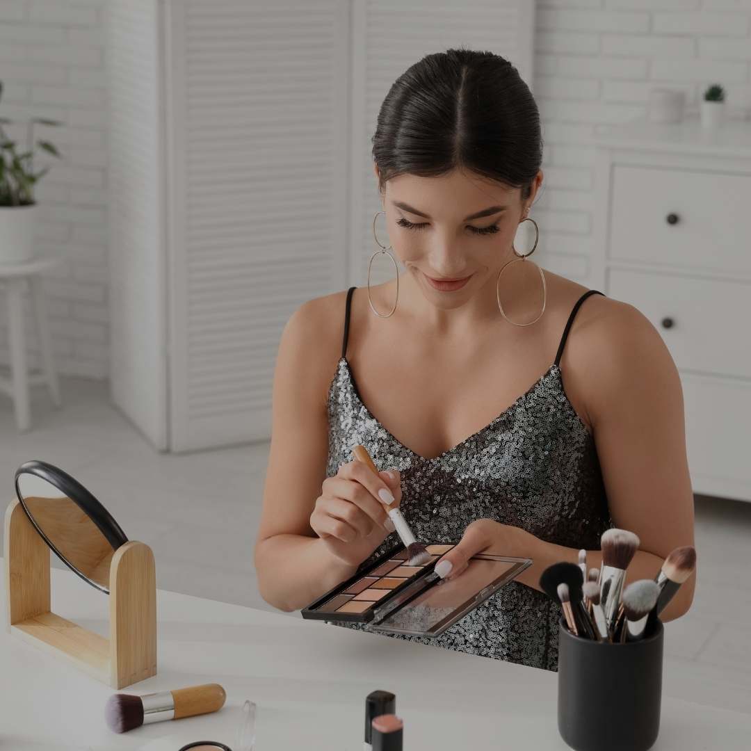 A woman with dark hair in a black sequined camisole sits at a white table, applying makeup from an eyeshadow palette with a brush. She is wearing large gold hoop earrings.