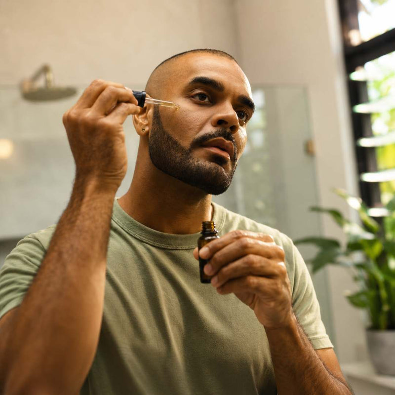 A man in a green t-shirt applies a drop of serum from a dropper bottle to his face in a bright bathroom setting.