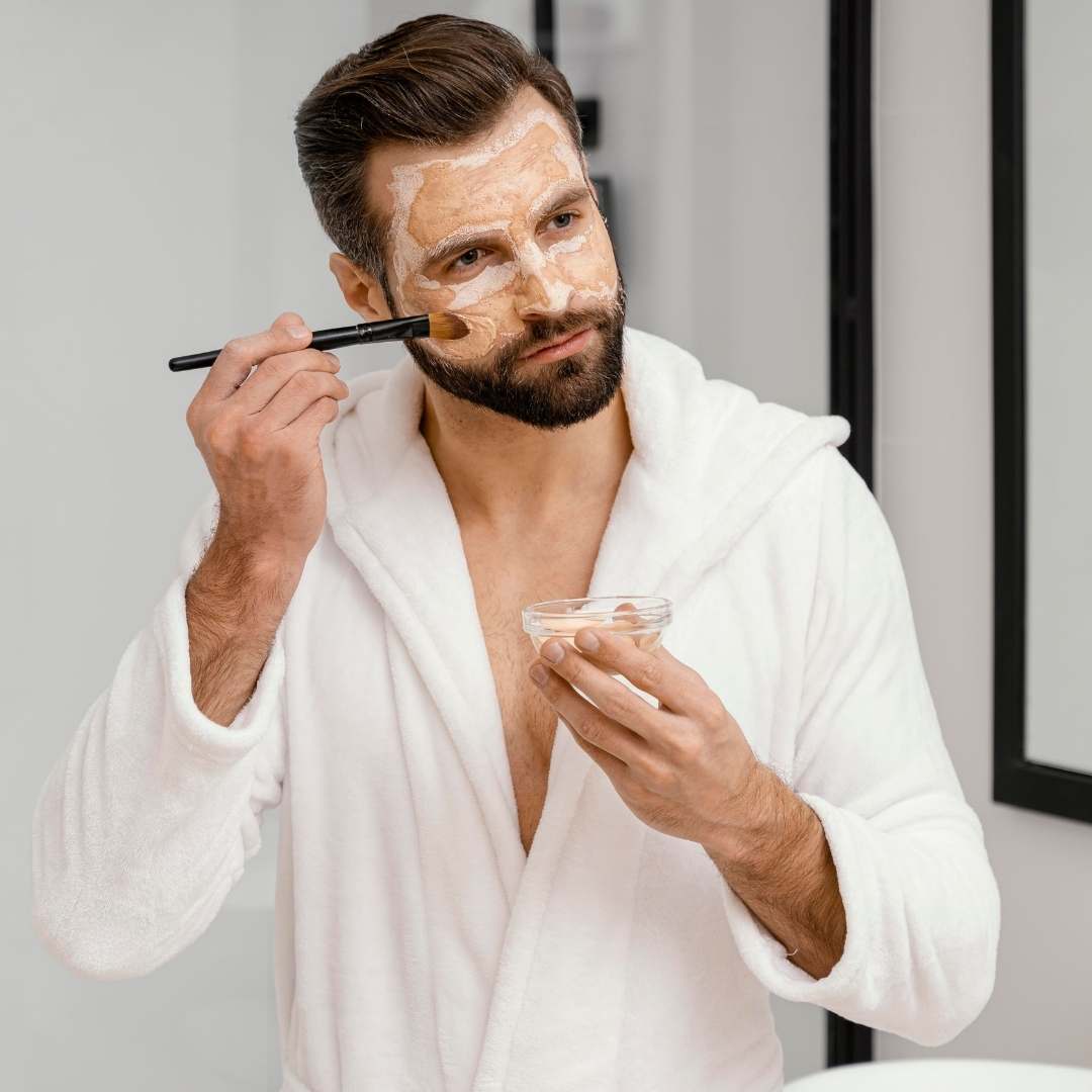 A man in a white bathrobe applies a beige facial mask or cream using a brush while holding a small clear bowl in a bright bathroom setting.