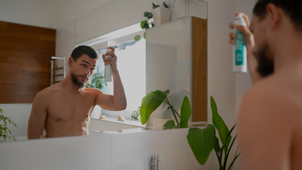 A man in a bathroom spraying a bottle of body wash or shower gel from a mirror, with his back and reflection visible. The scene includes a sink, wooden wall paneling, and green houseplants.