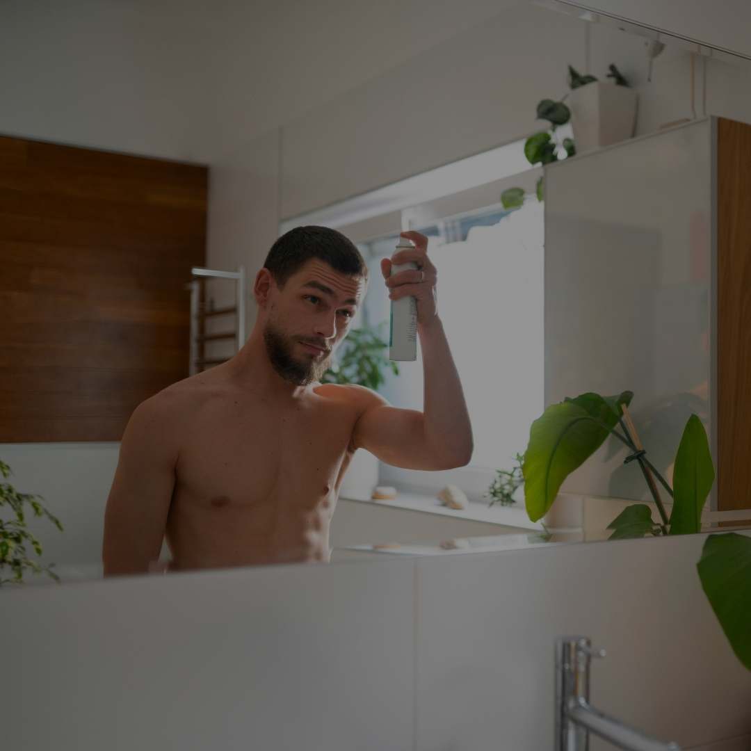A man in a bathroom spraying a bottle of body wash or shower gel from a mirror, with his back and reflection visible. The scene includes a sink, wooden wall paneling, and green houseplants.