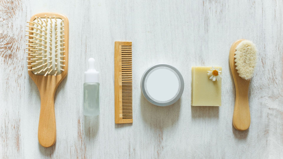 A flat-lay arrangement of natural hair and beauty care items on a white-washed wooden surface, including a wooden hairbrush, a small dropper bottle, a wooden comb, a round jar, a bar of soap with a daisy, and a small brush.