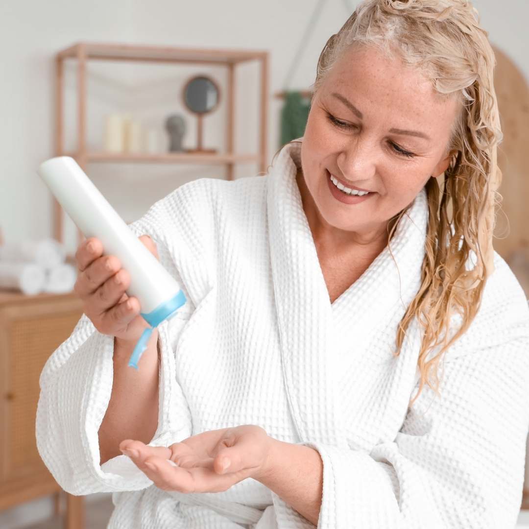 A smiling blonde woman in a white waffle robe holds a white bottle with a blue cap, preparing to dispense skincare cream into her hand in a bright bathroom setting.