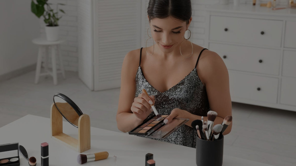 A woman in a sparkly black dress sits at a white vanity table, holding an eyeshadow palette and applying makeup with a brush. Makeup items, a small mirror, and a brush holder are arranged on the table in a bright, modern room.