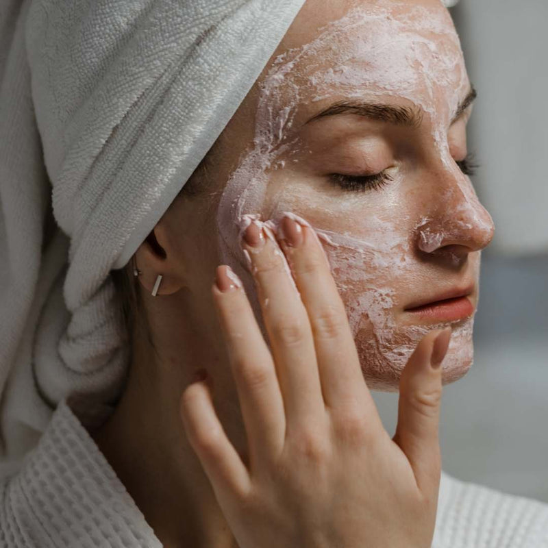 A close-up of a woman with a white towel wrapped around her hair, applying a white face mask cream to her cheek with her finger.