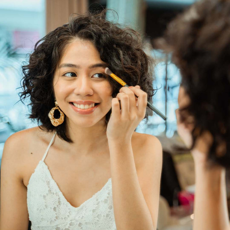 A woman with curly dark hair smiles while applying makeup with a brush to her eyebrow, wearing a white lace camisole and a large gold hoop earring.