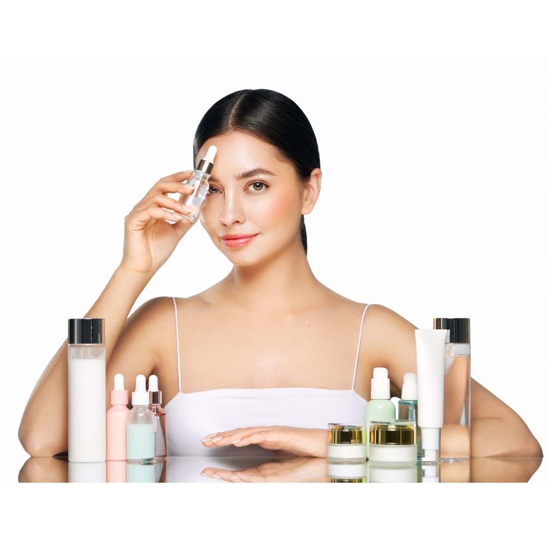 A woman with dark hair pulled back sits behind a table of skincare products, holding a clear dropper bottle near her face. The products include various bottles, jars, and tubes in white, pink, green, and gold packaging on a reflective white surface against a white background.