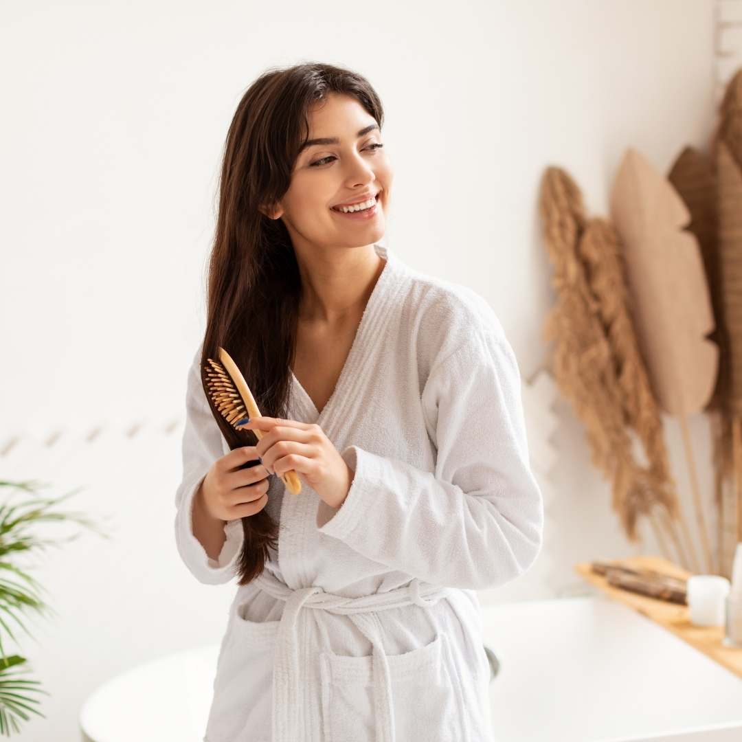 A woman with long dark brown hair wearing a white bathrobe holds a wooden hairbrush in a bright, modern bathroom setting.